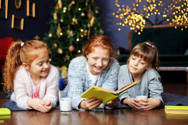 Three kids girls are reading a book on Christmas Eve. The concept of Merry Christmas, New Year, family and gifts. — Stock Photo, Image