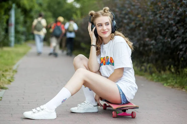 Jeune belle fille avec un skateboard. Écoutez de la musique sur votre casque. Le concept d'un mode de vie actif, passe-temps, musique . — Photo