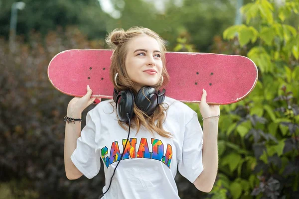 スケートボードを持つ10代の女の子。ヘッドフォンで音楽を聴く。アクティブなライフスタイル、趣味、音楽の概念. — ストック写真