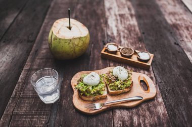 Tasty organic healthy breakfast - poached eggs on toast with avocado, water with lime, young coconut. Dark wooden background clipart