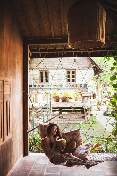 女人坐在吊床上 五颜六色的枕头和喝新鲜的小椰子 乡村风格的木屋屋 — 图库照片