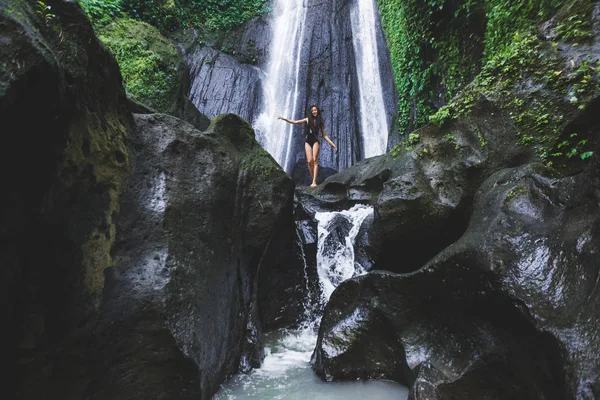 Woman enjoying near hidden in jungle cascade waterfall in Bali. Slim body and black swimsuit, fashion model. Dusun Kuning in Ubud area