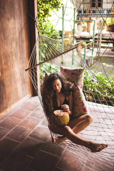 女人坐在吊床上 五颜六色的枕头和喝新鲜的小椰子 乡村风格的木屋屋 — 图库照片