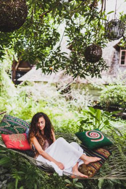 Bali bahçesinde hamak asılı rahatlatıcı beyaz elbiseli kadın. Ağaçta asılı yuvarlak ahşap hasır fener