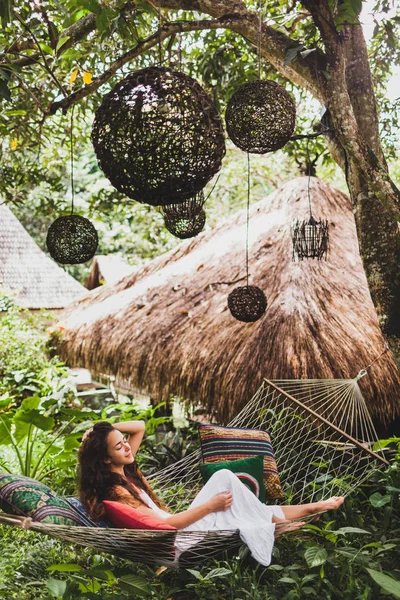 バリの庭でハンモックをぶら下げのリラックスした白のドレスを着た女性 木にぶら下がっている丸い木の枝編み細工品ランタン — ストック写真