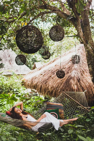 穿着白色衣服的女人在巴厘岛花园的吊床上放松 挂在树上的圆木柳条灯笼 — 图库照片