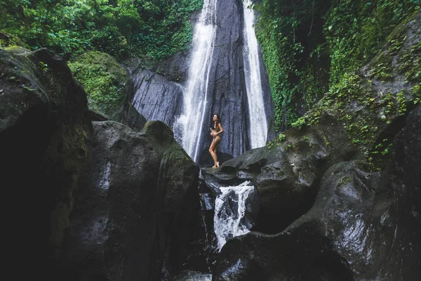 Woman enjoying near hidden in jungle cascade waterfall in Bali. Slim body and black swimsuit, fashion model. Dusun Kuning in Ubud area