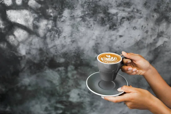 Šedý šálek čerstvého cappuccino v rukou ženy na pozadí gra — Stock fotografie