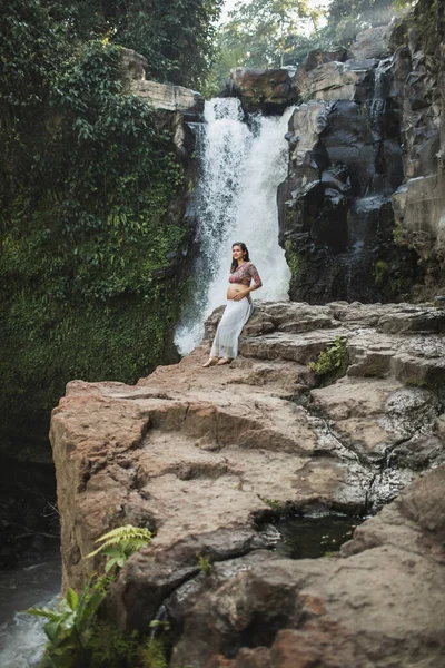 年轻的真正的孕妇接近惊人的瀑布瀑布 赤裸的腹部和白裙 与自然和谐相处 印度尼西亚Ubud Tegenungan — 图库照片