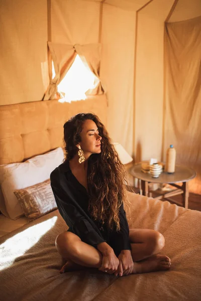 Sensual glamour portrait of oriental woman in interior of modern luxury glamping tent camp in Morocco. Relaxing on bed. Eco design concept, natural and wooden elements in style.