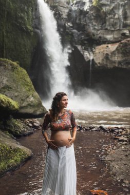 Genç, otantik hamile kadın şelalenin yanında. Çıplak göbek ve beyaz etek. Doğayla uyum. Gençlik, Ubud, Endonezya