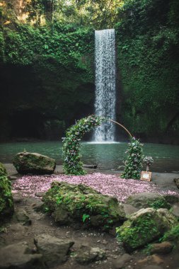 Yuvarlak bronz düğün kemeri pembe güller ve yeşilliklerle süslenmiş. Ormandaki küçük şelalenin yakınındaki tören için alışılmadık bir yer. Tibumana, Bali, Ubud.