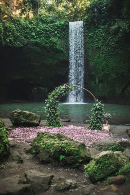Round bronze wedding arch decorated with pink roses and greens. Unusual location for ceremony near small waterfall in jungle. Tibumana, Bali, Ubud. clipart