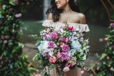 Gelin, düğün töreninde inanılmaz büyük bir çiçek buketi tutuyor. Pembe güller, şakayıklar, kırmızı laleler ve yeşiller yakın.