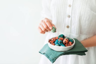 Paskalya için boyanmış bıldırcın yumurtalarıyla küçük güzel bir seramik tabak tutan kadın elleri. Yeşil peçete, beyaz kırsal elbise, kırmızı ve mavi boya. Yakın plan..