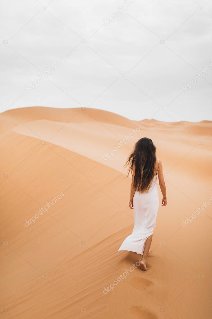 Woman with long hair in silk white dress walking in Sahara desert sand dunes in Morocco. View from the back.