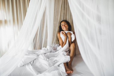 Güzel bronzlaşmış genç bir kadın beyaz yatakta uyanıyor. Mutlu uyanın ve yeni bir güne başlayın. Boş zaman ve dinlenme. Refah ve tasasız konsept. Uzun kıvırcık saçlı. Evde kal..