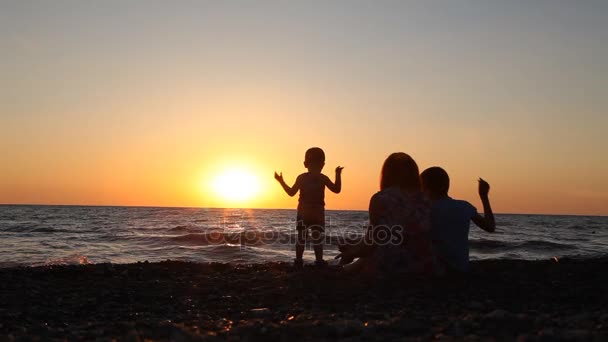 Silueta de mamá con dos niños sentados en una playa con hijos mar al atardecer — Vídeos de Stock