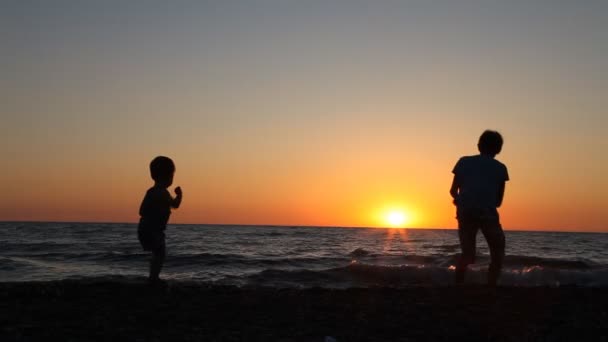 La silueta de dos chicos arrojando piedras al mar en la playa — Vídeo de stock