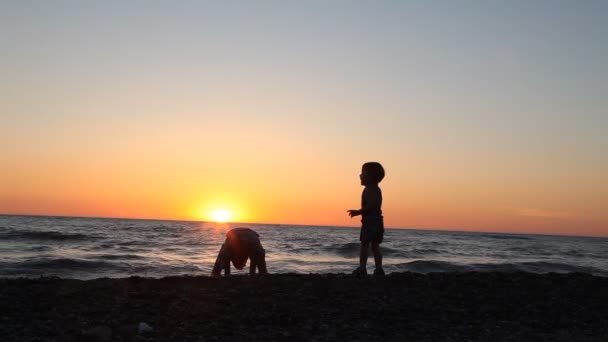 La silueta de dos chicos arrojando piedras al mar en la playa — Vídeos de Stock