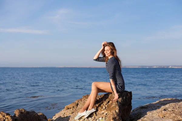 The girl in the striped dress sits on the rocks by the sea — Stock Photo, Image