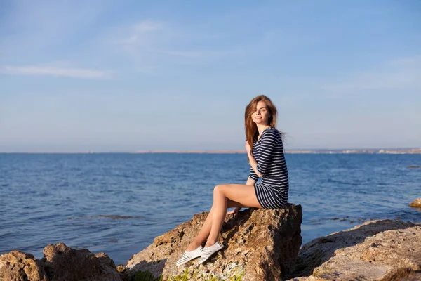 The girl in the striped dress sits on the rocks by the sea — Stock Photo, Image