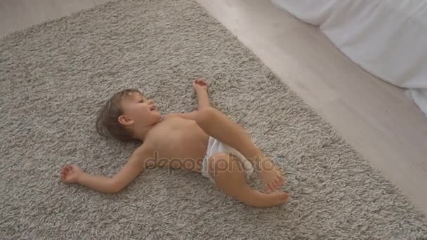 Маленький хлопчик лежить на килимі в спальні і грає — стокове відео