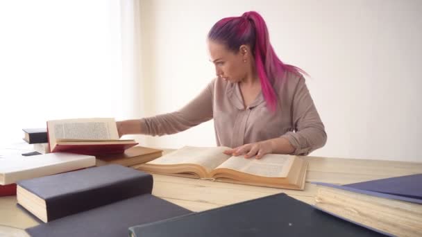 La chica con el pelo rosa está sentada en la mesa leyendo un libro — Vídeos de Stock