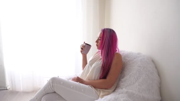 A menina com o cabelo rosa está sentada em uma poltrona branca bebendo café ou chá — Vídeo de Stock