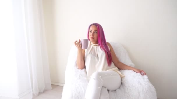 Das Mädchen mit den rosa Haaren sitzt in einem weißen Sessel und trinkt Kaffee oder Tee — Stockvideo
