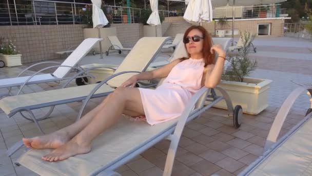 La chica con el pelo rojo broncea en el complejo junto al mar y la piscina — Vídeos de Stock