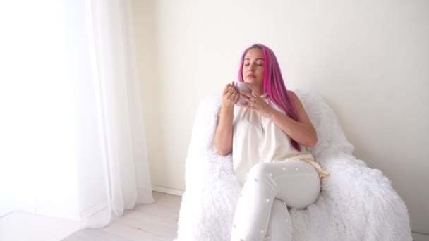 The girl with the pink hair is sitting in a white armchair drinking coffee or tea — Stock Video