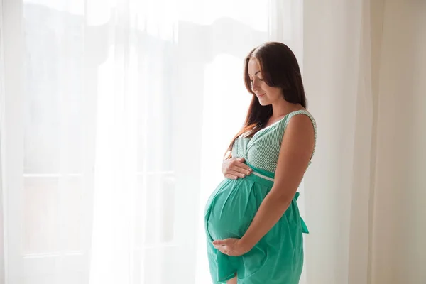 Pregnant woman in a white room stands at the window — Stock Photo, Image