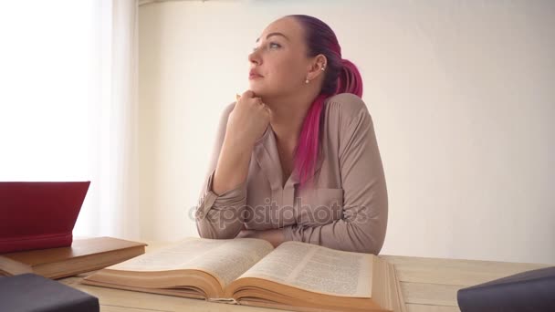 Девушка с розовыми волосами сидит за столом и читает книгу — стоковое видео