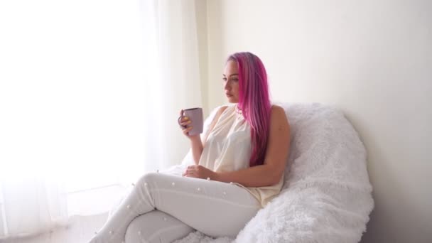 Η κοπέλα με το ροζ μαλλιά κάθεται σε ένα λευκό πολυθρόνα πίνοντας καφέ ή τσάι — Αρχείο Βίντεο
