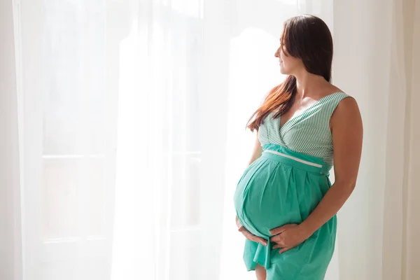 Zwangere vrouw in een witte kamer staat bij het raam — Stockfoto