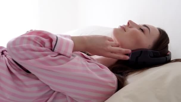 Schöne Frau im Pyjama lügt und hört Musik mit Kopfhörern — Stockvideo