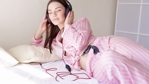 Kvinna i pyjamas på säng danserna till musik från hörlurar — Stockvideo