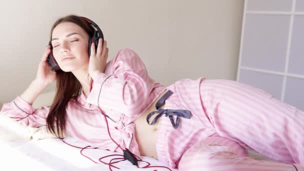 Vrouw in pyjama is liggend op het bed dansen op muziek van hoofdtelefoon — Stockvideo