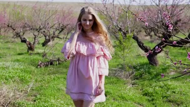 Блондинка в рожевій сукні гуляє квітучим садом з деревами — стокове відео
