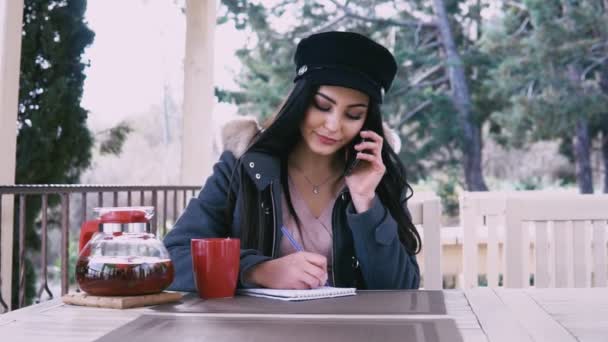 Schöne Brünette telefoniert mit heißem Tee im Straßenrestaurant — Stockvideo