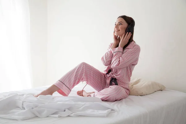 Brunette in roze pyjama's, luisteren naar muziek met een koptelefoon op het bed — Stockfoto