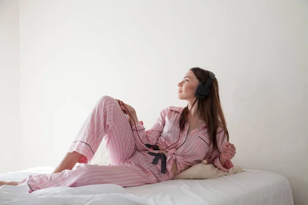 Брюнетка в розовом пижаме слушает музыку с наушниками на кровати — стоковое фото