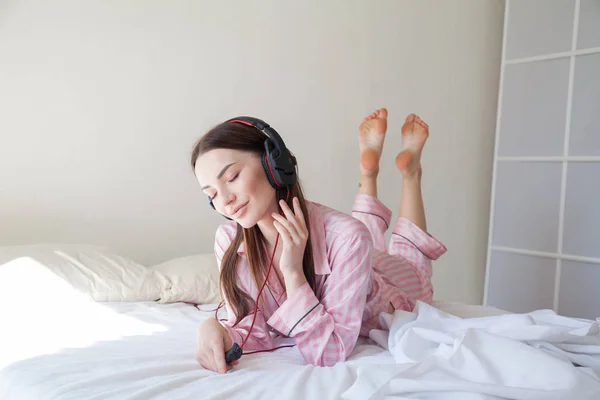 Brunetka v růžovém pyžamku poslechu hudby se sluchátky na posteli — Stock fotografie