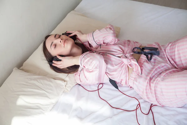 Девушка в розовом пижаме лежит на кровати и слушает музыку с наушниками — стоковое фото