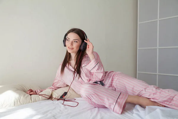 Bruna in pigiama rosa ascoltare musica con le cuffie sul letto — Foto Stock