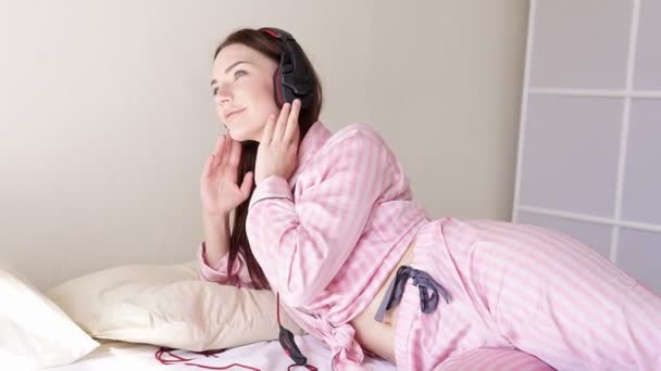 Женщина в пижаме на кровати танцует под музыку из наушников — стоковое видео