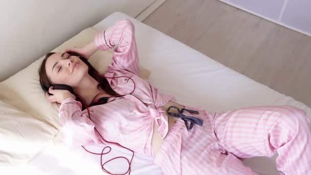 Жінка в піжамі лежить на ліжку танцює до музики з навушників — стокове відео