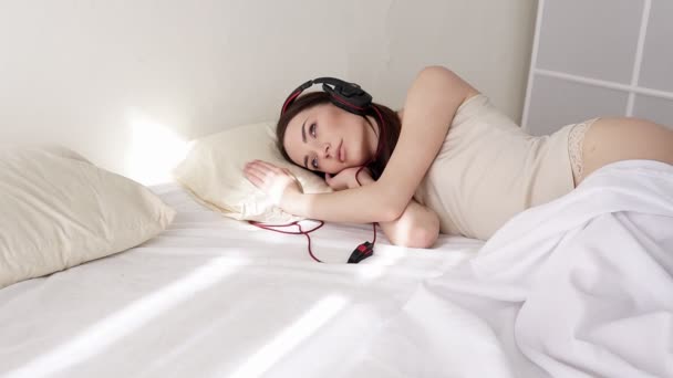 Woman in lingerie lying in the bedroom listening to music from big headphone — Stock Video