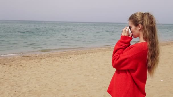 सुनहरे बालों वाली लड़की समुद्र के किनारे समुद्र पर चाय पीती है — स्टॉक वीडियो
